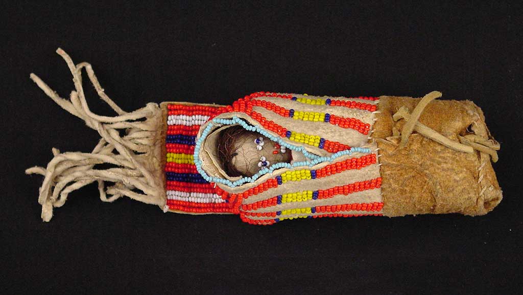 small doll with tiny blue eyes wrapped up in a blanket decorated with red, yellow, and blue beadwork